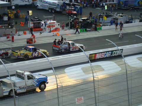 1995 Ford Bronco - Race trucks are on the track with support vehicles and pit crews in the background at a NASCAR event.