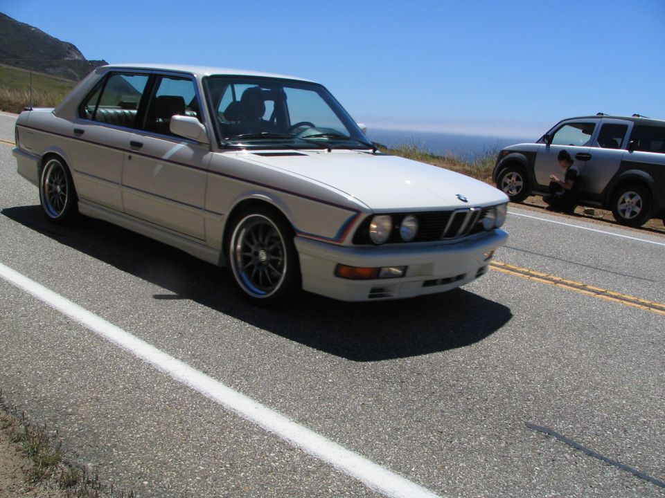 White BMW sedan with red and blue pinstripes, driving on a coastal road
