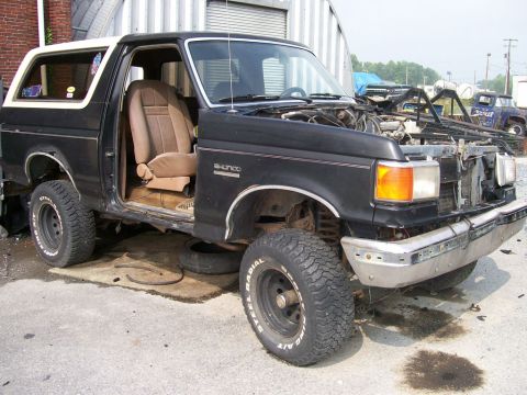 1989 Ford Bronco - A black Ford Bronco with a white top sits in a lot, missing its hood and driver's door, with mechanical parts visible.