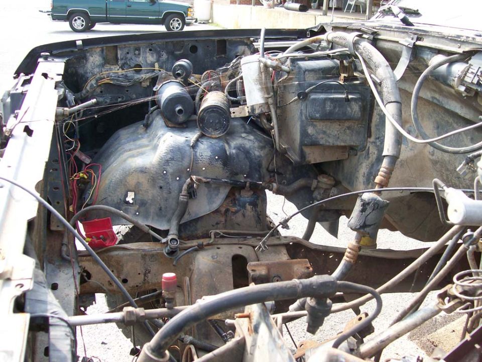 Engine bay of a damaged vehicle, showing hoses, wires, and engine components. A green pickup truck is visible in the background