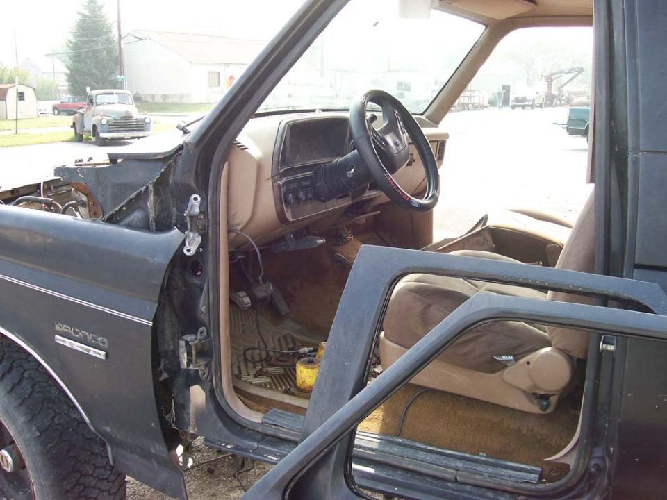 Dark gray 1980s Ford Bronco interior with tan seats and dashboard, driver's door open, showing exterior and another truck in background