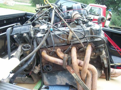 1989 Ford Bronco - Engine components are visible in the bed of a truck, with a red truck and two people in the background.