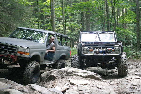 Two modified Ford Broncos navigating a rocky forest trail. The front Bronco has a winch and roll cage