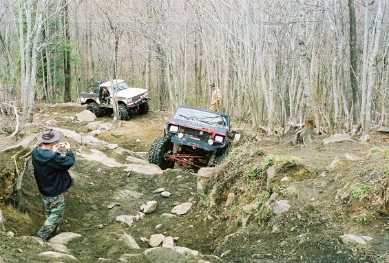 Two off-road vehicles on a muddy, wooded trail; one is climbing a steep incline