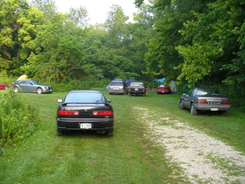 1991 Ford Probe - Several vehicles are parked in a grassy area surrounded by trees, with tents set up in the background.