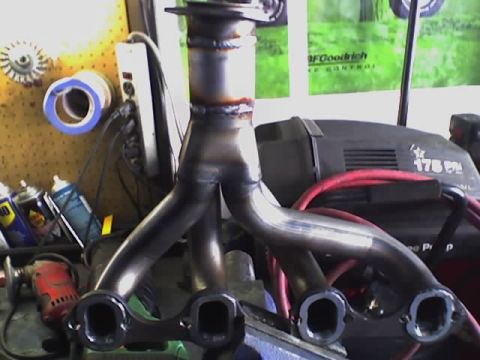 1971 Ford Bronco - A custom exhaust manifold is clamped in a vise, with tools and equipment in the background.