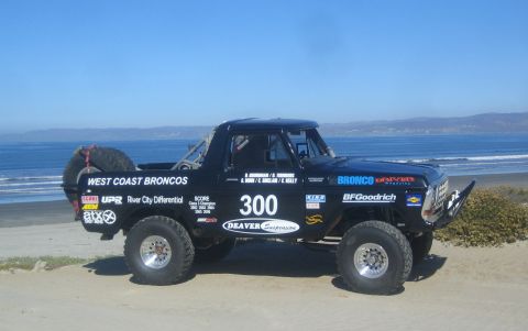 1990 Ford Bronco - black tube-chassis off-road race truck numbered 300 with West Coast Broncos and BFGoodrich livery on a beach.