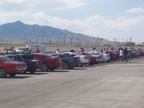 1972 Ford Ranchero - A row of cars are parked on an asphalt lot with mountains visible in the distance under a partly cloudy sky.