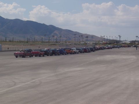 1972 Ford Ranchero - A long row of vehicles are parked on a paved surface with a mountain range visible in the distance under a cloudy sky.