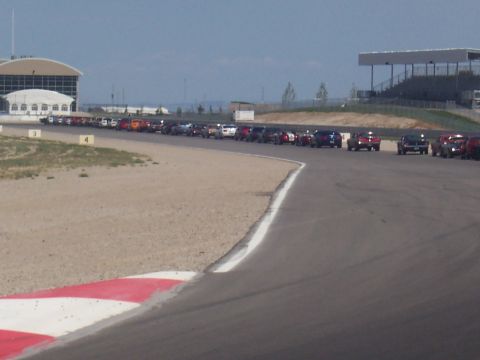 1972 Ford Ranchero - A long line of vehicles is parked on the side of a racetrack, with grandstands visible in the distance.