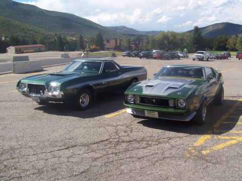 1972 Ford Ranchero - Two classic American muscle cars, a Ford Ranchero and a Ford Mustang, are parked side-by-side in a lot with mountains in the background.
