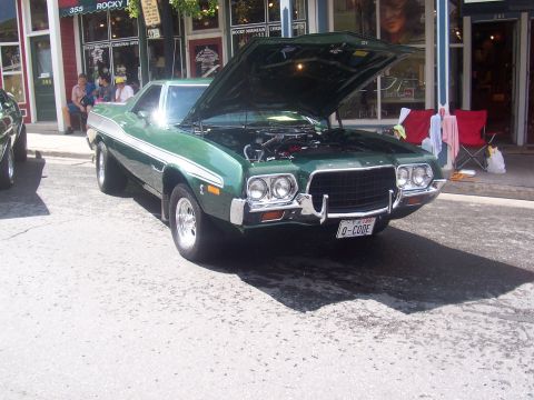 1972 Ford Ranchero - A green 1972 Ford Ranchero is displayed with its hood open, parked in front of shops.