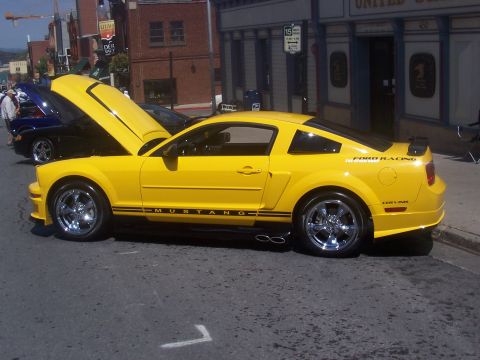 1972 Ford Ranchero - A bright yellow Ford Mustang is parked on a street with its hood open, displaying black racing stripes and "Ford Racing" badging.