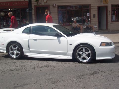 1972 Ford Ranchero - A white Ford Mustang GT is parked on a street in front of shops with people sitting outside.