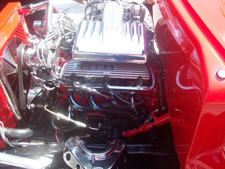 Engine bay showing chrome Cobra valve covers, red block, and fan. Some red bodywork visible