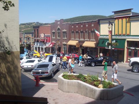 1972 Ford Ranchero - A silver muscle car with black hood stripes is parked on a street lined with shops, alongside other classic vehicles.