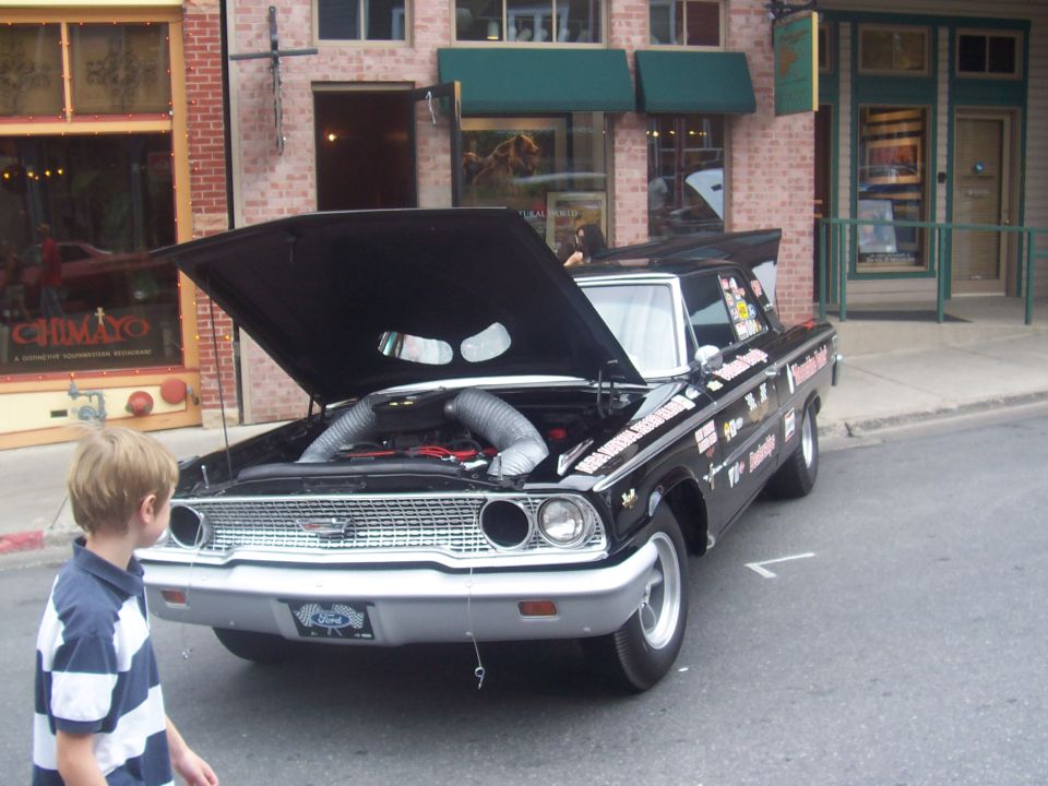 Black Ford sedan with hood open, showing engine and intake tubes. Covered in racing decals
