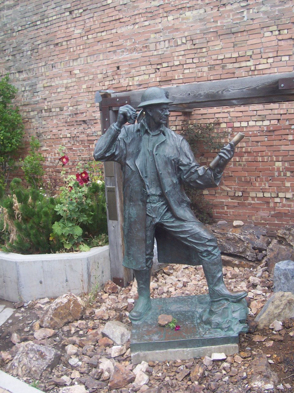 Bronze statue of a man wearing a trench coat and hat, holding a scroll or tube, standing outdoors