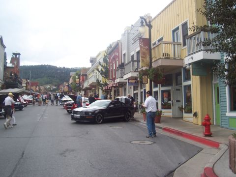 1972 Ford Ranchero - A black Ford Mustang is parked on a street lined with shops, with other vehicles and pedestrians present.