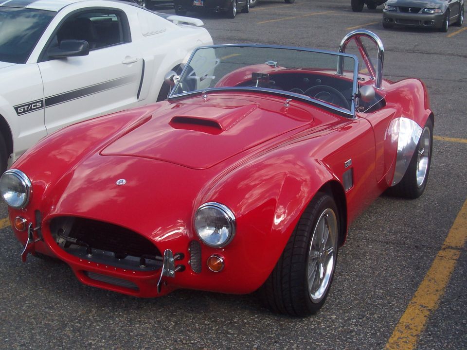 Red sports car with a roll bar and scoop on the hood. White Ford Mustang GT/CS parked behind it
