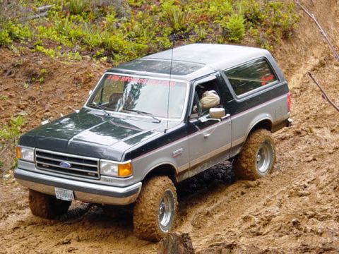 1989 Ford Bronco - This two-tone Ford Bronco navigates a challenging muddy incline, with visible red text on the windshield.