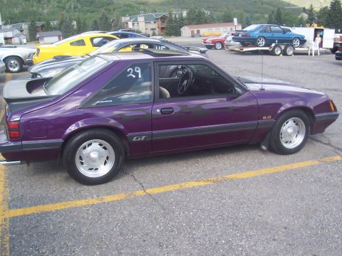 1972 Ford Ranchero - A purple 1987 Ford Mustang GT is parked in a lot with other vehicles and buildings in the background.