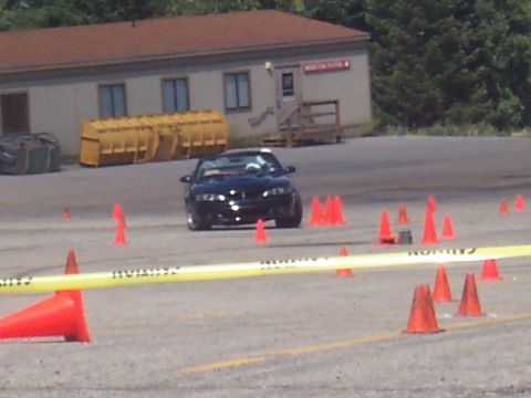 1972 Ford Ranchero - A black sports car drives through a course set up with cones and caution tape in front of a building.