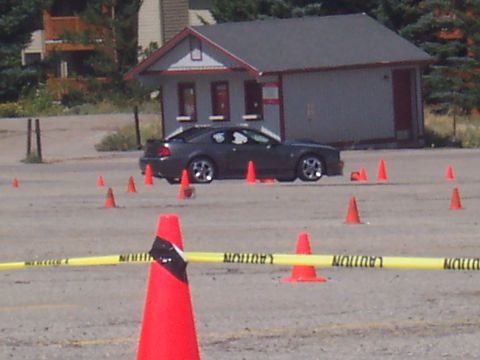 1972 Ford Ranchero - A dark coupe drives through an obstacle course marked by cones and caution tape in front of a small building.
