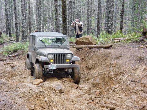 1989 Ford Bronco - A Jeep Wrangler navigates a challenging, muddy off-road trail surrounded by trees.
