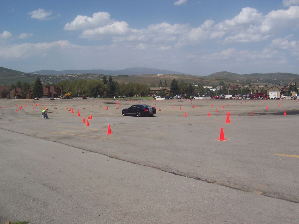 Black car navigating a cone course in a large paved lot with buildings and hills in the background