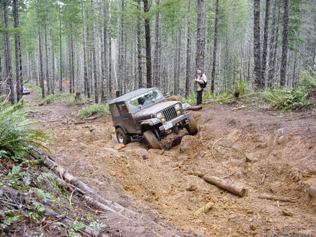 Tan Jeep Wrangler YJ soft top crawling through a muddy rut on a forest trail