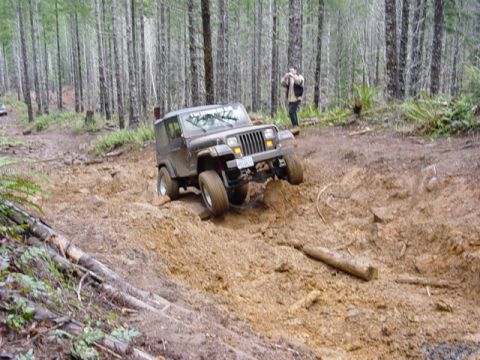 1989 Ford Bronco - A Jeep Wrangler tackles a challenging muddy trail in a wooded area, with a person observing in the background.