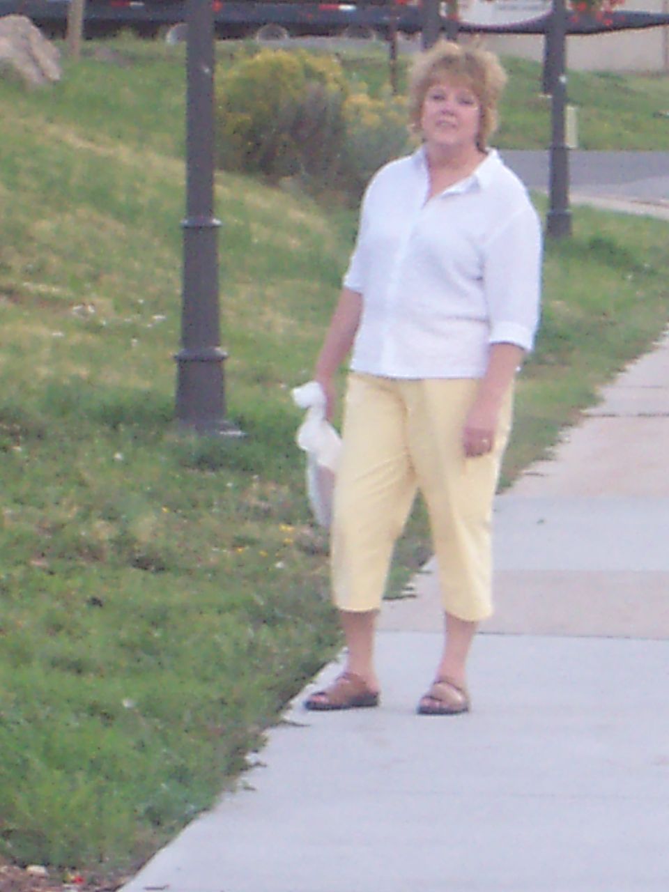 Woman in a white shirt and yellow capri pants stands on a sidewalk next to a grassy area