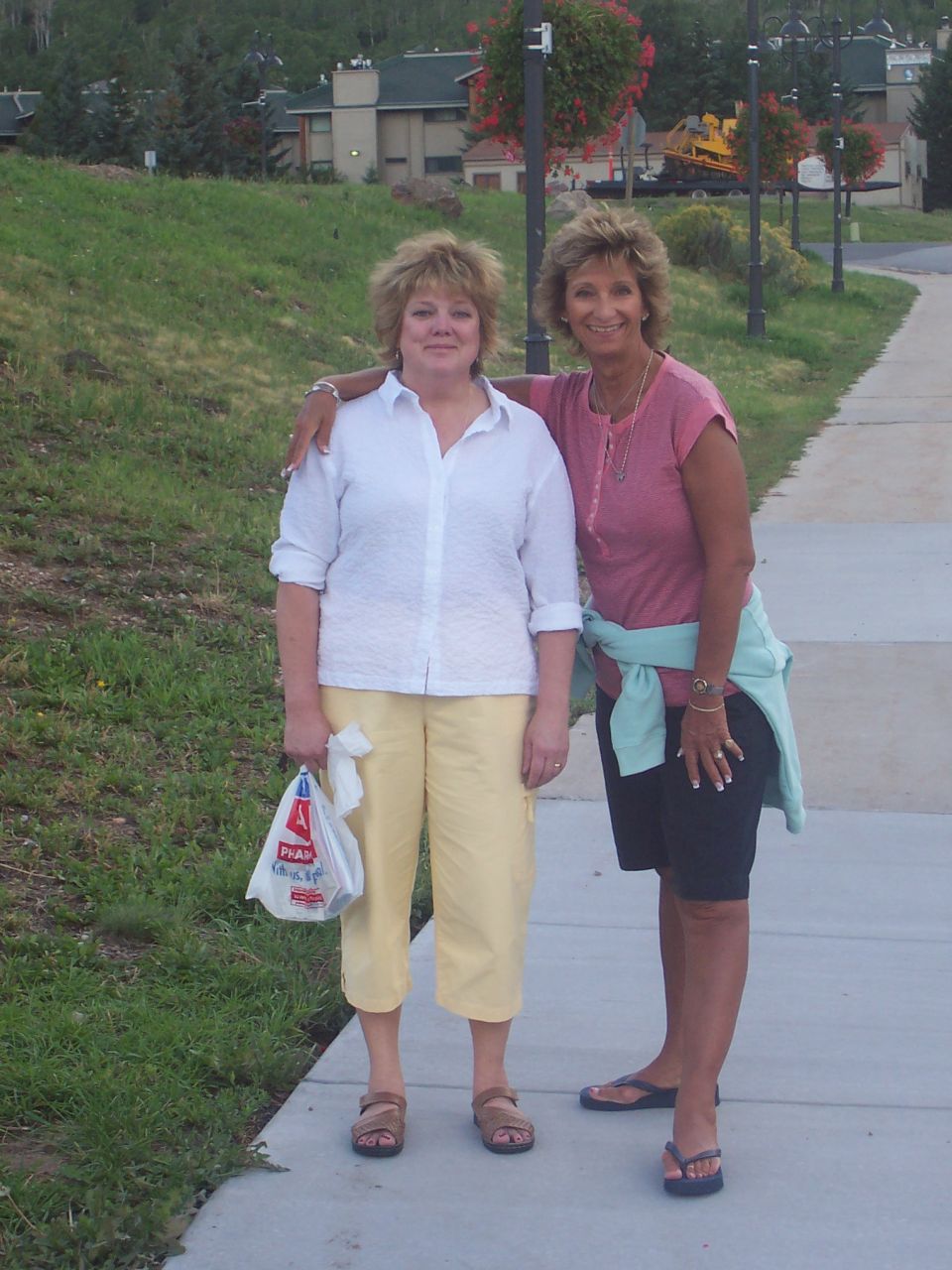 Two women stand on a sidewalk with buildings and trees in the background