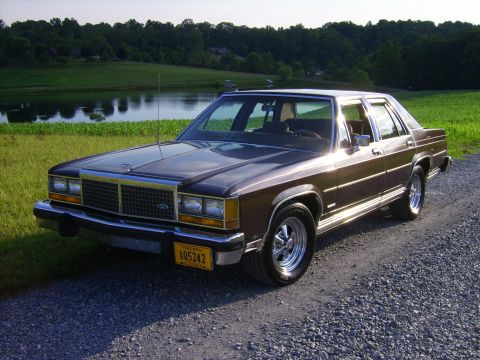 1982 Ford LTD Crown Victoria - Front three-quarter view of a dark brown Ford LTD Crown Victoria four-door sedan parked on a gravel drive above a farm pond, with tan cloth…