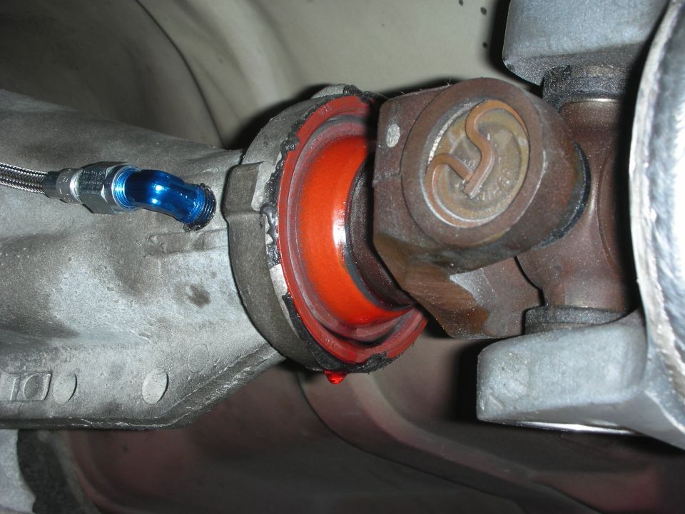 Close-up of a driveshaft U-joint connected to a transmission mount with a red rubber bushing. A blue fitting with a braided line is attached to the transmission housing