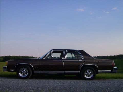 1982 Ford LTD Crown Victoria - Full side profile of a dark brown Ford LTD Crown Victoria four-door sedan on a gravel drive at dusk, with an open field and treeline behind…