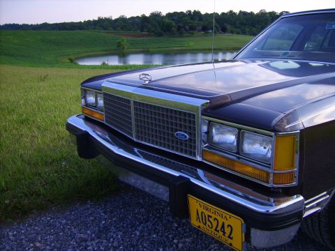 1982 Ford LTD Crown Victoria - Tight low-angle detail of the front end of a dark brown Ford LTD Crown Victoria sedan on a gravel drive above a farm pond, showing the brig…