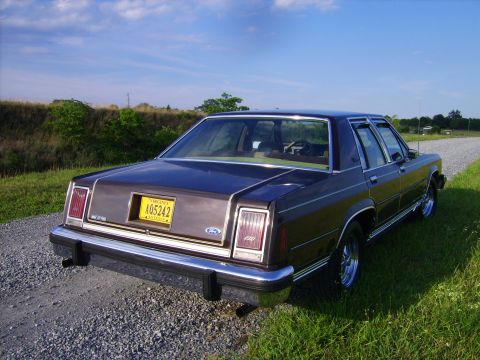 1982 Ford LTD Crown Victoria - Three-quarter rear view of a dark burgundy/maroon 1980s Ford LTD Crown Victoria four-door sedan parked on a gravel shoulder beside a grassy…