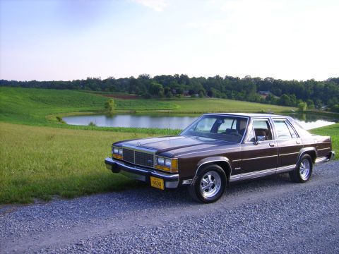 1982 Ford LTD Crown Victoria - A brown 1980 Ford LTD Crown Victoria is parked on a gravel road next to a pond with rolling green hills in the background.