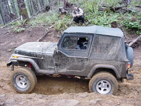 1989 Ford Bronco - A Jeep Wrangler is stuck in thick mud on a wooded trail, with a person observing from the embankment above.