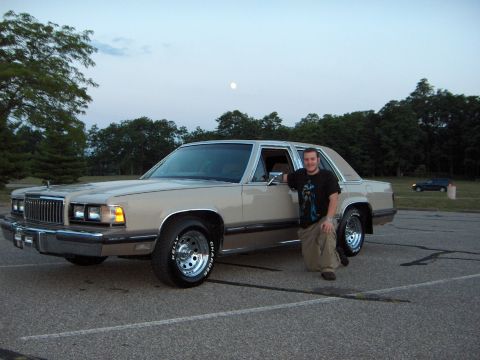 1988 Mercury Grand Marquis - A man poses next to a tan 1988 Mercury Grand Marquis in a parking lot with trees and a distant car in the background.