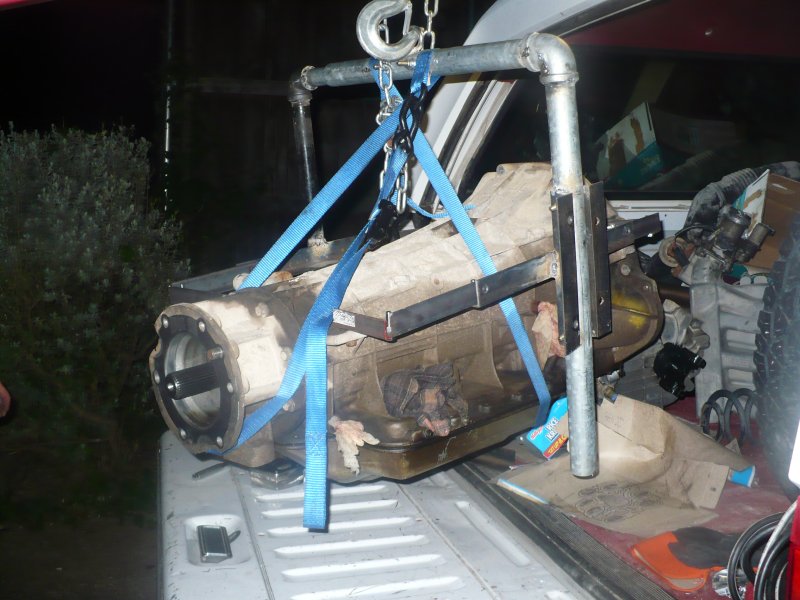Automatic transmission hanging from blue straps and chain hoist on pipe frame in a truck bed