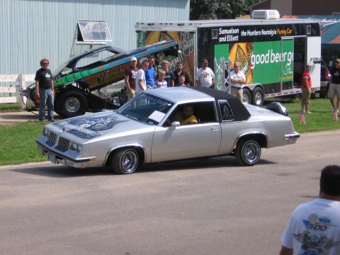 1989 Ford Mustang - A silver coupe with a custom graphic on the hood is displayed at an outdoor event, with a dragster and trailer in the background.