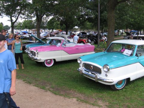 1989 Ford Mustang - A pair of vintage Nash Metropolitan cars are displayed at a car show with other vehicles in the background.