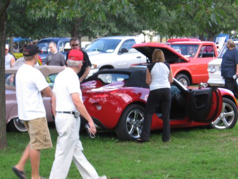 1989 Ford Mustang - A red convertible is displayed at an outdoor car show with several other vehicles and spectators present.