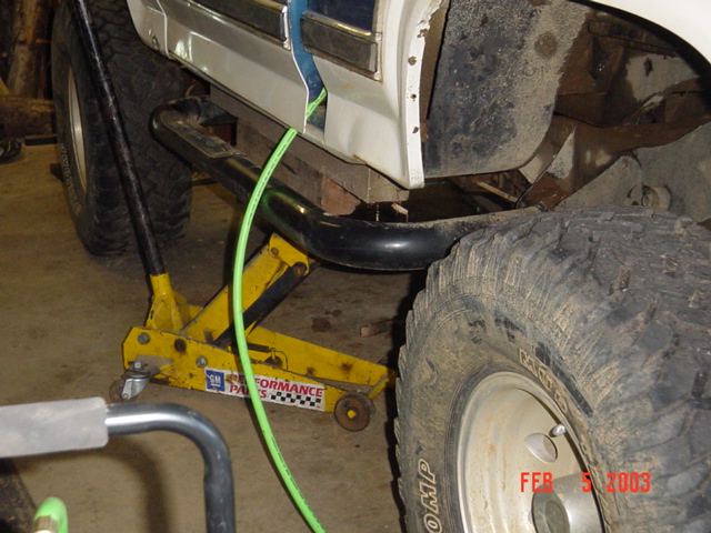 Floor jack under the rocker of a white-and-blue truck in a garage, with green hose
