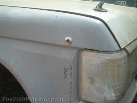 1990 Ford Bronco - Front fender corner of a dirty white truck showing a hood pin, clear marker light and a handwritten inventory label.