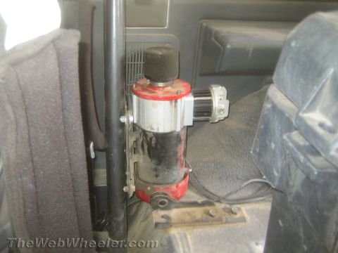 1990 Ford Bronco - red and polished metal hydraulic lever unit bolted to the floor next to a seat inside a dusty vehicle interior.