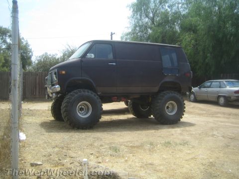 1968 Ford Mustang - A heavily modified van with oversized tires sits in a dirt lot, with another car visible in the background.